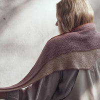 An image of a person facing a white wall. Draped across their shoulders is a shawl in shades of cream and pale purple with orange along the top edge.