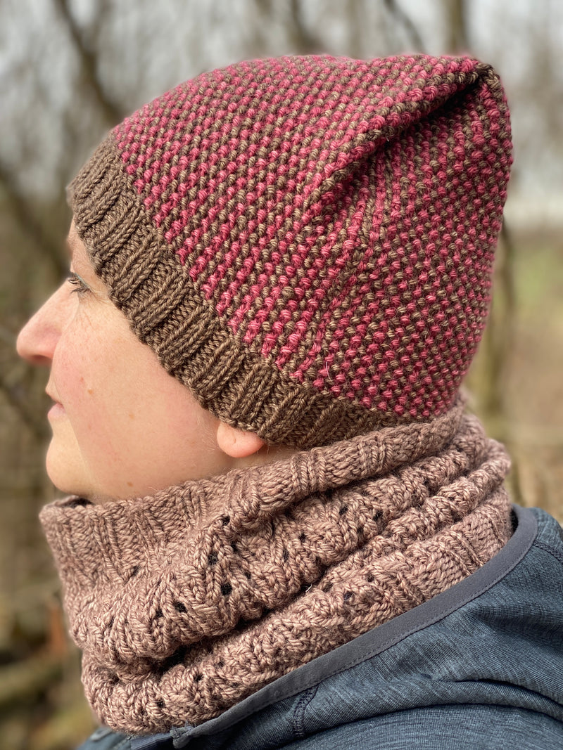 A side profile image of Victoria wearing a brown cowl and a red and brown hat. The knitted hat has a brown ribbed brim and the body has vertical stripes alternating red and brown. The crown has a folded pleat on the visible side