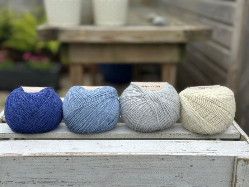 Four balls of wool in royal blue, medium denim blue, silvery grey, and cream.