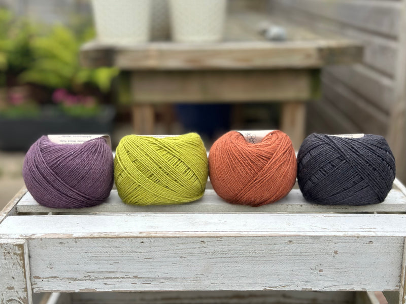 Four balls of wool in dark aubergine purple, earthy lime green, rust, and dark grey.