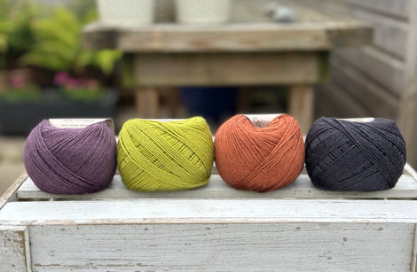 Four balls of wool in dark aubergine purple, earthy lime green, rust, and dark grey.