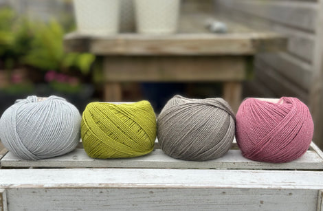 Four balls of wool in silvery grey, rich earthy lime green, medium grey, and medium soft pink.