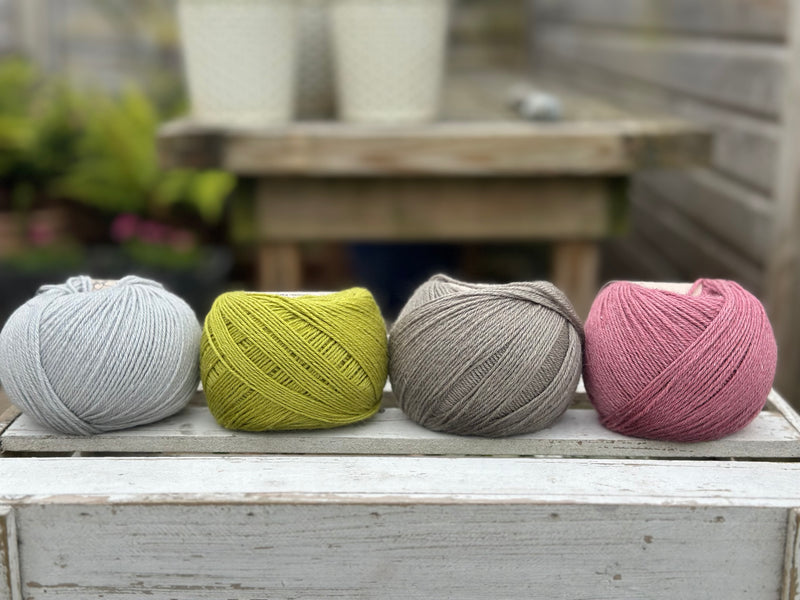Four balls of wool in silvery grey, rich earthy lime green, medium grey, and medium soft pink.