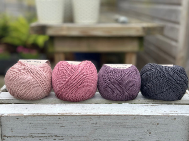 Four balls of wool in light warm pink, medium soft pink, dark aubergine purple, and dark grey.