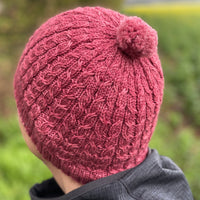 A person wearing a pink knitted hat is facing away from the camera. The hat has a cable pattern and a small bobble on the crown