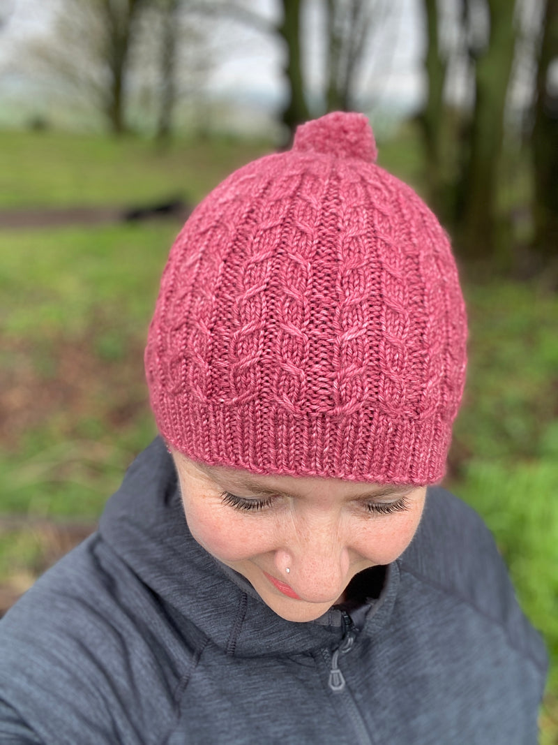 Victoria looking towards the floor wearing a pink knitted hat. The hat has a cable pattern and a small bobble on the crown