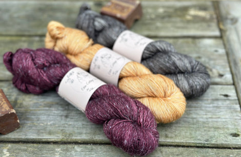 Three skeins of yarn. From left to right: a dark purple skein, a golden brown skein and a dark grey skein