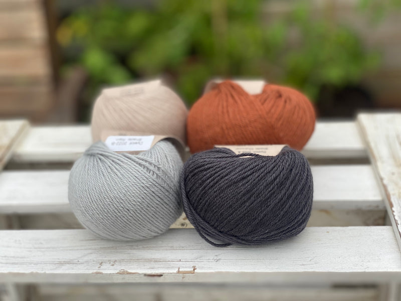 Four balls of yarn in two rows of two balls. On the left there is a beige ball and a pale blue ball. On the right there is a reddish brown ball and a dark grey ball