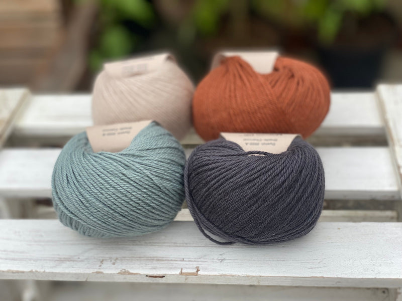 Four balls of yarn in two rows of two balls. On the left there is a beige ball and a blue-green ball. On the right there is a reddish brown ball and a dark grey ball