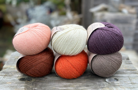 Six balls of yarn arranged in two rows of three balls. The top row is peachy orange, cream and dark purple. The bottom row is reddish brown, orange and grey