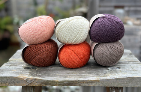 Six balls of yarn arranged in two rows of three balls. The top row is peachy orange, cream and dark purple. The bottom row is reddish brown, orange and grey