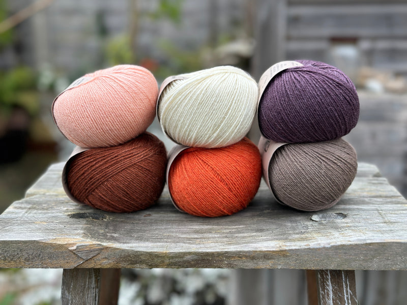 Six balls of yarn arranged in two rows of three balls. The top row is peachy orange, cream and dark purple. The bottom row is reddish brown, orange and grey