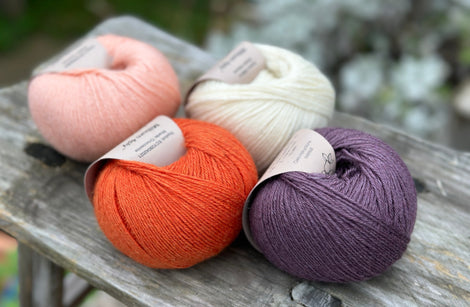 Four balls of yarn. From left to right there is a peachy pink ball, an orange ball, a cream ball and a dark purple ball.