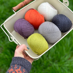 Six balls of yarn in a white metal bowl. The colours are dark purple, orange, cream, bright green, grey and black