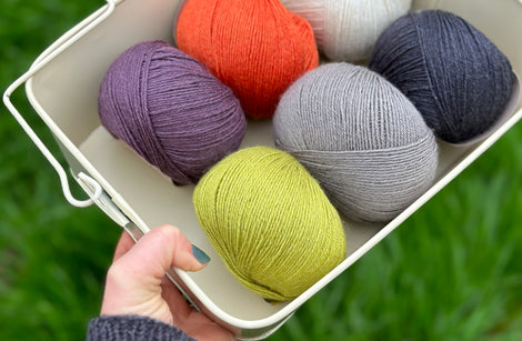 Six balls of yarn in a white metal bowl. The colours are dark purple, orange, cream, bright green, grey and black