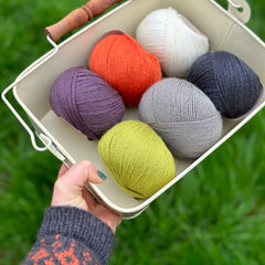 Six balls of yarn in a white metal bowl. The colours are dark purple, orange, cream, bright green, grey and black