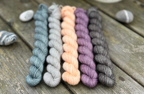 Five mini skeins of yarn - from left to right - a blue skein, a grey skein, a peachy orange skein, a purple skein and a dark grey skein