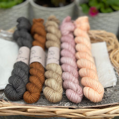 Five mini skeins of yarn. From left to right - a black skein, a reddish-brown skein, a pale brown skein, a pink skein with gold sparkle running through it and a peach skein