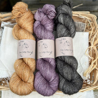 Three skeins of yarn. From left to right; a golden brown skein, a purple skein and a dark grey skein