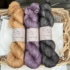 Three skeins of yarn. From left to right; a golden brown skein, a purple skein and a dark grey skein