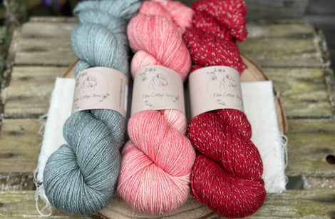 Three skeins of yarn with silver sparkle running through it. From left to right: a grey skein, a pink skein and a red skein