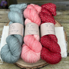 Three skeins of yarn with silver sparkle running through it. From left to right: a grey skein, a pink skein and a red skein