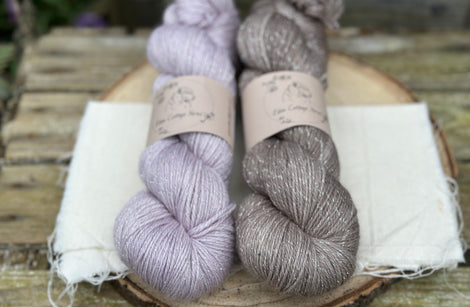 Two skeins of yarn with silver sparkle running through it. One pale purple skein and one mid brown skein