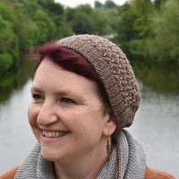 Victoria facing to the left of the camera in front of a lake. She is wearing a brown knitted hat with a lace pattern and beads.