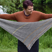 Victoria holding a triangular grey shawl outstretched. The shawl is grey with thin stripes of contrasting colours