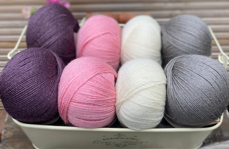 Eight balls of yarn in four pairs. From left to right the colourways are purple, pink, cream and grey