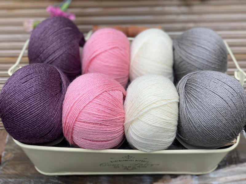 Eight balls of yarn in four pairs. From left to right the colourways are purple, pink, cream and grey