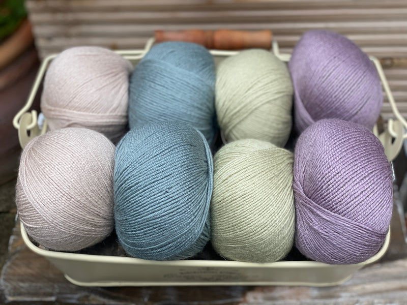 Eight balls of yarn in four pairs. From left to right the colourways are beige, blue-green, pale green, light purple
