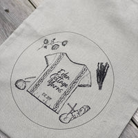 A rectangular pale grey cotton drawstring bag. A circular logo is printed on the bag. The logo has a cable design tee with Eden Cottage Yarns Est 2011 on it, a flower stem, a bunch of herb-like shrubbery, a ball of yarn with a crochet hook and a skein of yarn with knitting needles.