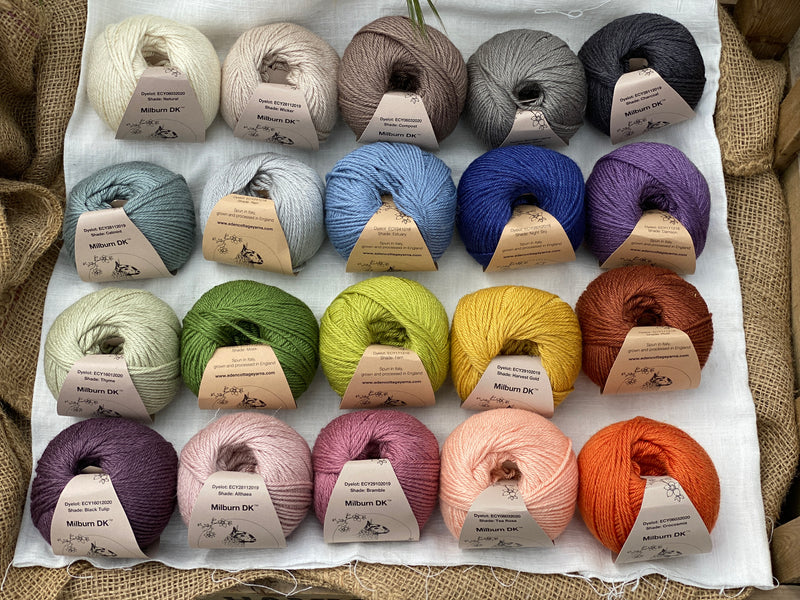 Twenty balls of yarn are arranged in four rows of five balls. The yarn is resting on a white piece of fabric in a jute lined wooden box. From left to right, the colours in the top row are Natural, Wicker, Compost, Steel and Charcoal. The colours in the second row are Catmint, Rain, Estuary, Night Sky and Damson. The colours in the third row are Thyme, Moss, Fern, Harvest Gold and Rust. The colours in the bottom row are Black Tulip, Althaea, Bramble, Tea Rose and Crocosmia.