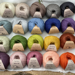 Twenty balls of yarn are arranged in four rows of five balls. The yarn is resting on a white piece of fabric in a jute lined wooden box. From left to right, the colours in the top row are Natural, Wicker, Compost, Steel and Charcoal. The colours in the second row are Catmint, Rain, Estuary, Night Sky and Damson. The colours in the third row are Thyme, Moss, Fern, Harvest Gold and Rust. The colours in the bottom row are Black Tulip, Althaea, Bramble, Tea Rose and Crocosmia.