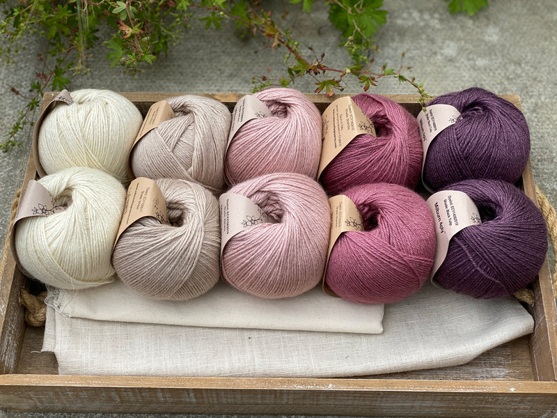 10 balls of yarn are sat in a wooden tray, there are two balls of each colour. The colours from left to right are Natural, Wicker, Althaea, Bramble and Black Tulip. The yarns create a fade effect from natural cream, through pink to dusky purple.