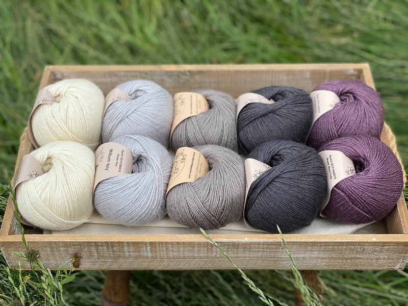 10 balls of yarn are sat in a wooden tray surrounded by grass. There are two balls of each colour. The colours from left to right are Natural, Rain, Steel, Charcoal and Black Tulip. The yarns create a fade effect from natural cream, through shades of grey ending with an accent of dusky purple.