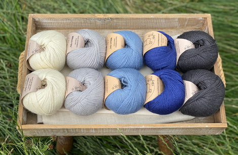 10 balls of yarn are sat in a wooden tray surrounded by grass. There are two balls of each colour. The colours from left to right are Natural, Rain, Estuary, Night Sky and Charcoal. The yarns create a fade effect from natural cream through shades of blue, ending in dark grey.