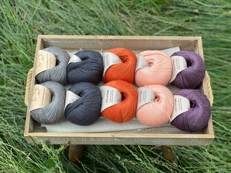 10 balls of yarn are sat in a wooden tray surrounded by grass. There are two balls of each colour. The colours from left to right are Steel, Charcoal, Crocosmia, Tea Rose and Black Tulip. The yarns create a palette of greys with orange, peach and dusky purple.