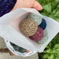 A glassine bag containing Yarnlings is held in the palm of a hand. The bag contains five DK weight Yarnlings in a lucky dip of colours. This bag has brown, purple, dark blue and pale green yarns.