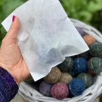 A glassine bag containing Yarnlings is held in the palm of a hand. The feint outline of Yarnlings can be seen through the bag. In the background a white wicker basket containing Yarnlings in a variety of colours can be seen.