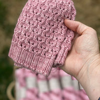 A pink knitted hat is folded and held in front of a blurry background. The background has a basket of pink skeins of yarn
