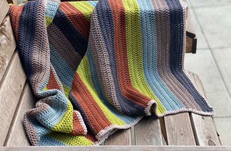 A striped crochet blanket draped over a bench. The stripes are rust, green, blue, brown and black and the blanket has a beige edging