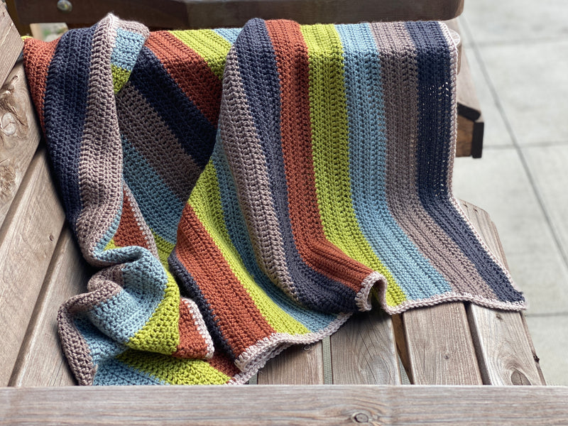 A striped crochet blanket draped over a bench. The stripes are rust, green, blue, brown and black and the blanket has a beige edging