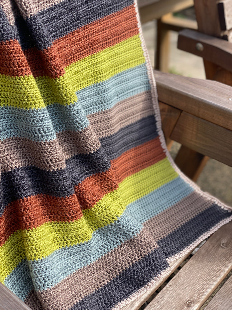 A striped crochet blanket draped over a bench. The stripes are rust, green, blue, brown and black and the blanket has a beige edging