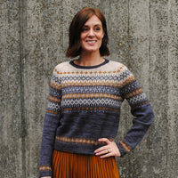 Rachel stood in front of a wall wearing a colourwork jumper with intricate detailing to the yoke, sleeves and body. The colours in the jumper are cream, light grey, brown and grey.