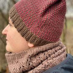 A side profile image of Victoria wearing a brown cowl and a red and brown hat. The knitted hat has a brown ribbed brim and the body has vertical stripes alternating red and brown. The crown has a folded pleat on the visible side