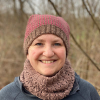 Victoria smiling at the camera wearing a brown cowl and a knitted hat. The knitted hat has a brown ribbed brim and the body has vertical stripes alternating red and brown. The crown of the hat has a peak on each side of the head