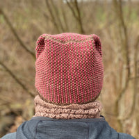 A person facing away from the camera wearing a grey jacket, a brown cowl and a knitted hat. The knitted hat has vertical stripes alternating red and brown. The crown of the hat has folded peaks on either side of the head