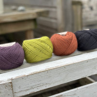 Four balls of wool in dark aubergine purple, earthy lime green, rust, and dark grey. 